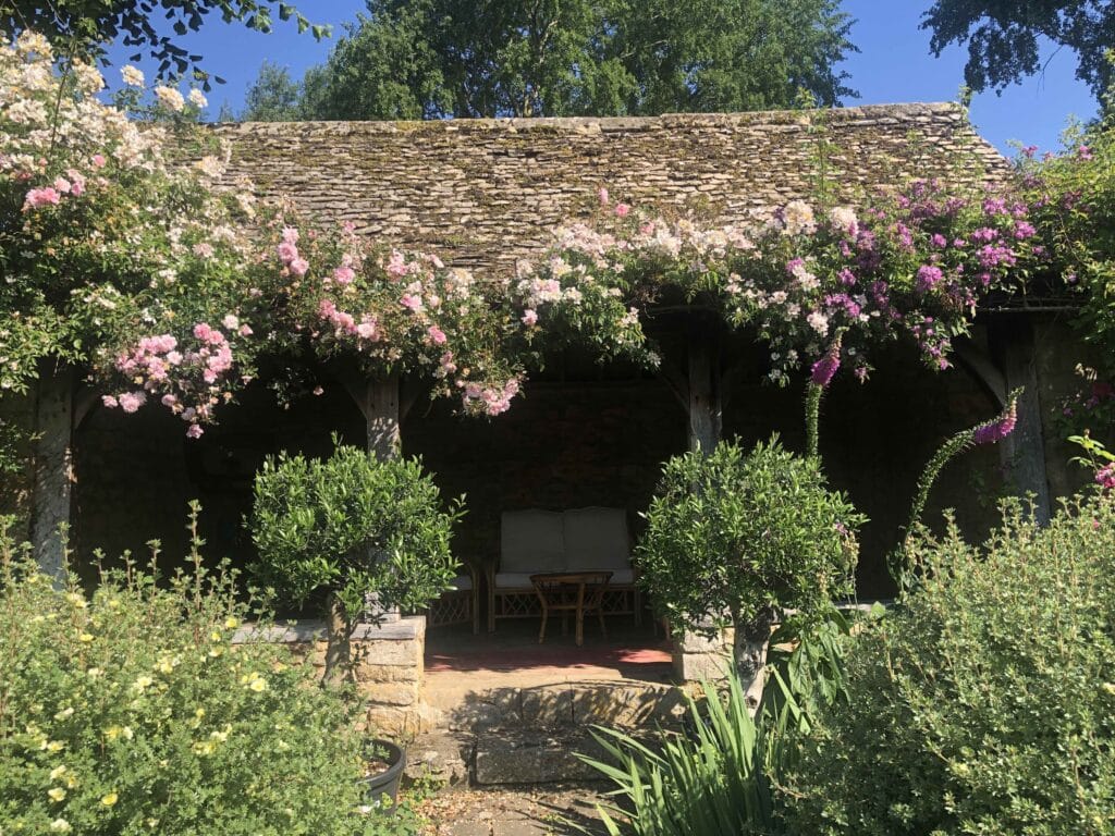 A rustic stone patio shaded by a wooden pergola, covered in pink and white climbing roses, offers the charm of luxury travel. Two green bushes frame the entrance, leading to a cozy seating area with a chair and table. Lush greenery surrounds the scene under a clear blue sky. David Pearson Travel