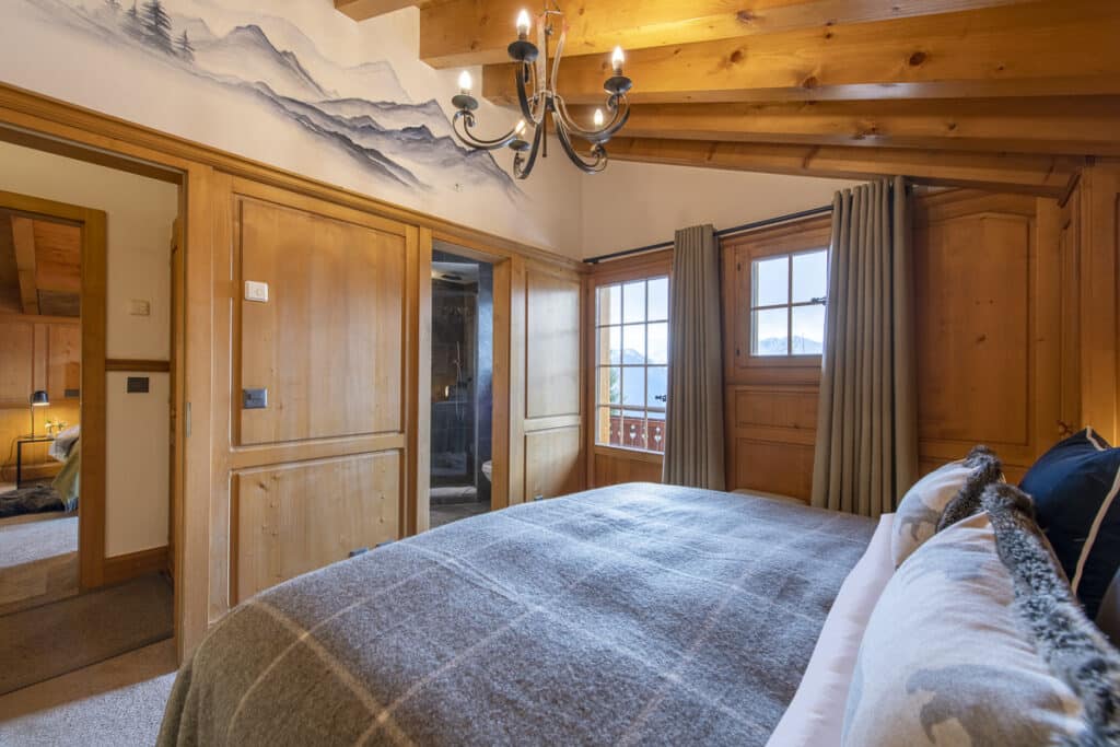 Cozy bedroom with wooden walls and ceiling, featuring a soft bed with plaid bedding. A chandelier adds a touch of luxury. Large windows with curtains let in natural light, offering a view outside. A door on the left leads to another room, perfect for those who appreciate refined travel comforts. David Pearson Travel