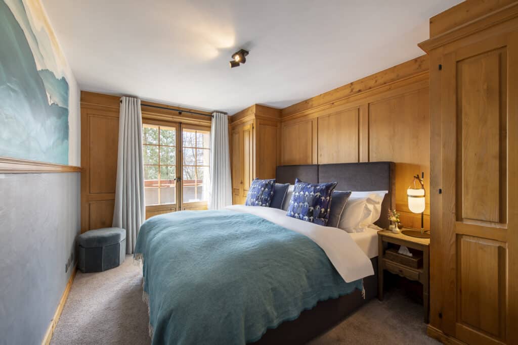 A cozy bedroom with wood-paneled walls offers a hint of luxury travel. The large bed features a blue throw and patterned pillows, complemented by a small nightstand with a lamp. Natural light flows through the windows light curtains, illuminating a round ottoman to the side. David Pearson Travel