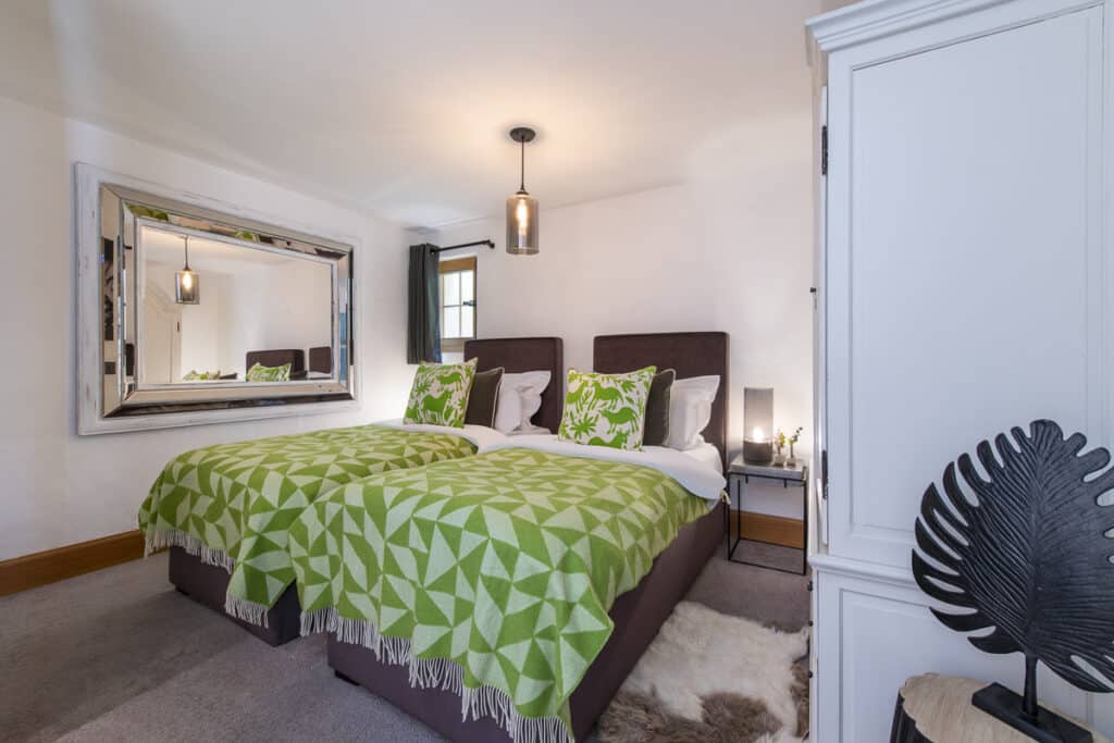 A cozy bedroom perfect for luxury travel, featuring two beds draped in green geometric-patterned blankets with matching pillows. A large mirror graces the wall, reflecting the elegance. A soft rug adds comfort underfoot, while a pendant light casts a warm glow. A sleek white wardrobe completes the space. David Pearson Travel