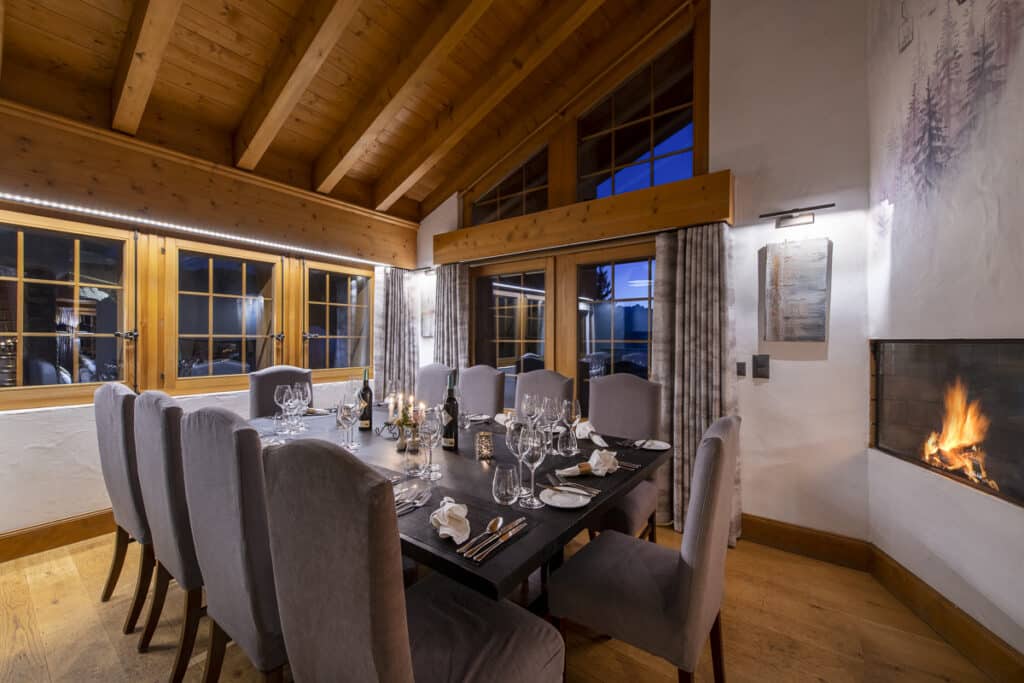 A cozy dining room exudes the charm of luxury travel, featuring a long table set for a meal and surrounded by gray upholstered chairs. With large windows, a wooden ceiling, and a lit fireplace, it offers an inviting atmosphere reminiscent of an elegant getaway. David Pearson Travel