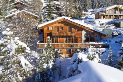 A cozy wooden chalet, ideal for luxury travel, is surrounded by snow-covered trees and hills in a winter landscape. With a stone foundation, large windows, and balconies, it stands amidst other similar houses. Snow blankets the roof and surroundings, creating a serene escape. David Pearson Travel