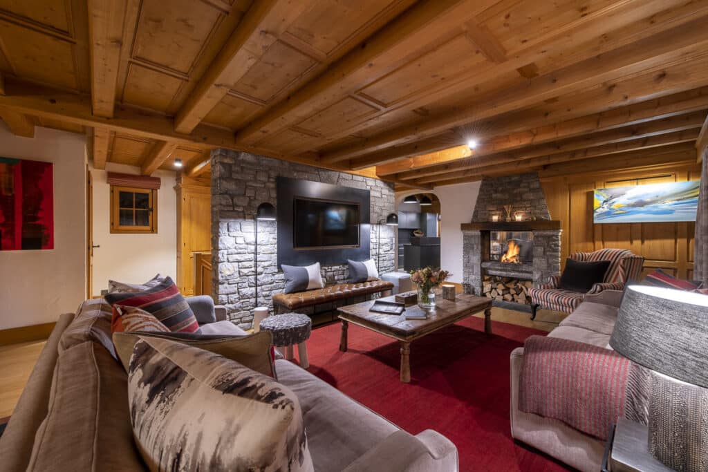 Cozy living room with wooden beam ceiling, stone fireplace, and modern furnishings reminiscent of luxury travel. A large TV is mounted on a stone wall. Sofas and armchairs with cushions surround a wooden coffee table on a red rug. Warm lighting enhances the welcoming ambiance.
. David Pearson Travel