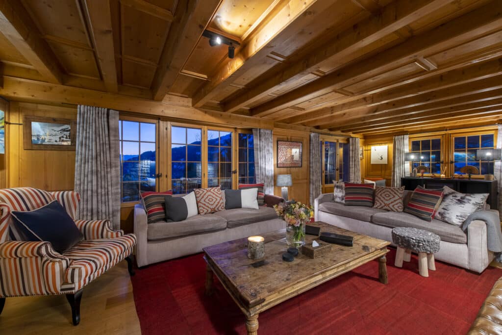 Cozy living room with two sofas and armchairs, wooden ceiling and walls, large windows revealing a snowy mountain view. A rustic coffee table sits on a red rug, adorned with flowers. Soft lighting creates a warm atmosphere, offering a touch of luxury travel right at home. David Pearson Travel