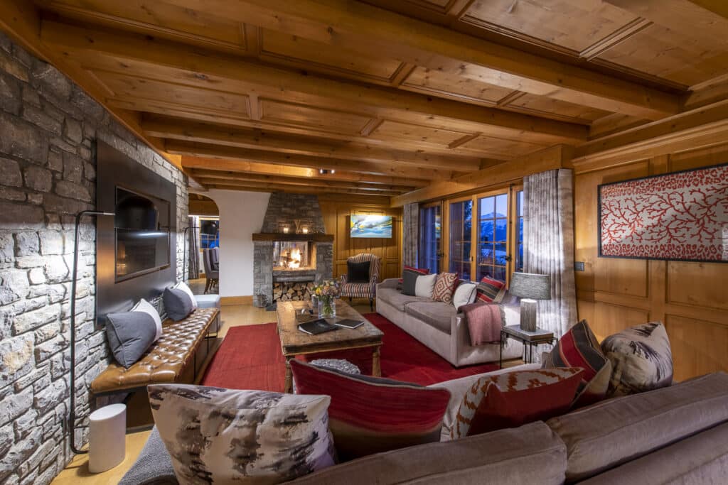 Cozy living room with wooden ceiling and stone walls exudes a touch of luxury travel. Features a fireplace, modern TV, and plush seating with colorful pillows. A coffee table and a patterned red rug are in the center. Large windows reveal an enchanting evening view. David Pearson Travel