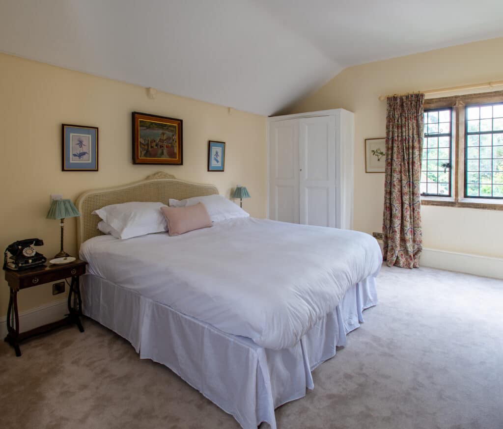 A cozy bedroom perfect for luxury travel, featuring cream-colored walls and a large bed with crisp white linens. Two lamps with blue shades sit on small bedside tables, complementing the white wardrobe. Floral curtains reveal lush greenery, while framed art adds an elegant touch. David Pearson Travel