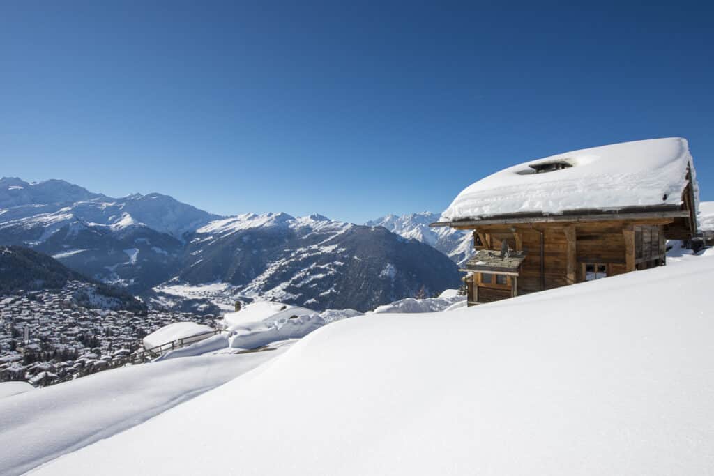 A wooden cabin with a snow-covered roof sits on a snow-blanketed hillside, offering a luxury travel experience as it overlooks a valley and distant snow-capped mountains under a clear blue sky. David Pearson Travel