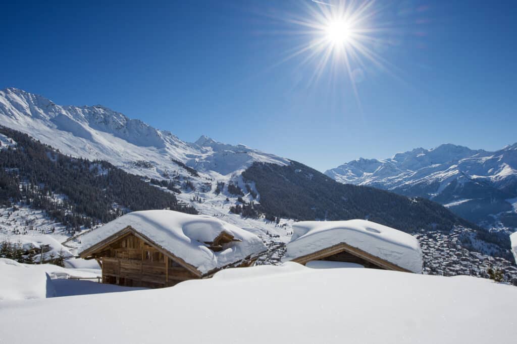 Snow-covered wooden cabins in the foreground offer a luxury travel experience with panoramic views of snowy mountains under a clear blue sky. The sun shines brightly, casting light on a mountainous landscape adorned with evergreen forests. David Pearson Travel