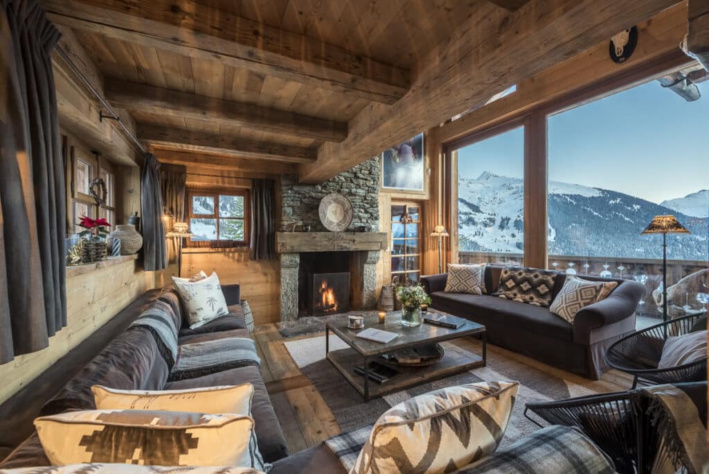 Cozy wooden chalet living room with large windows showcasing a snowy mountain view. This epitome of luxury travel features a stone fireplace, plush sofas with patterned cushions, a rug, and rustic decor, creating a warm and inviting atmosphere. David Pearson Travel