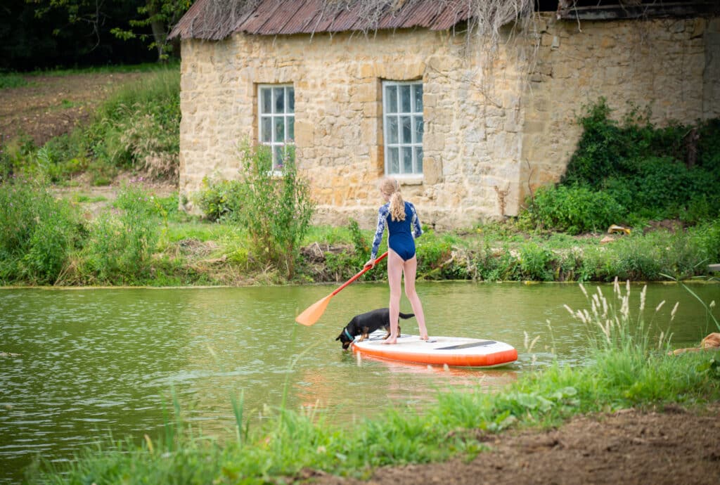 A person with blonde hair, in a blue swimsuit, gracefully paddles a sleek paddleboard alongside their black dog on a serene green pond. Near the rustic stone building with two windows and a tin roof, this idyllic scene captures the essence of luxury travel amidst lush greenery. David Pearson Travel