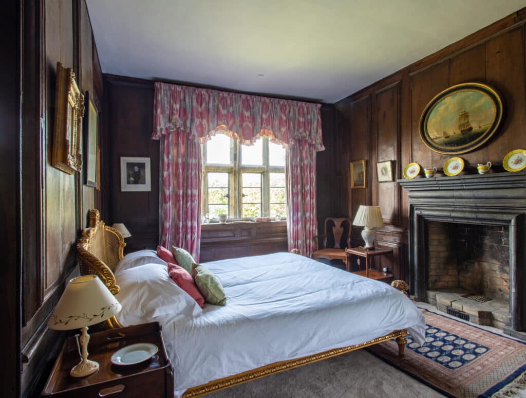 A cozy, vintage bedroom with wood-paneled walls, an ornate fireplace, and a large window adorned with pink curtains offers a touch of luxury travel allure. The bed, dressed in white linens and decorative pillows, is flanked by wooden nightstands with lamps. Vintage decor accents the room. David Pearson Travel