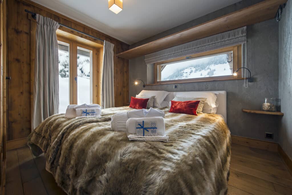 Cozy bedroom with a large bed covered in a furry blanket and luxury travel-inspired folded towels featuring blue embroidery. Red cushions accent the bed, while a snowy landscape is visible through the window. Wooden walls and flooring create a warm ambiance. David Pearson Travel
