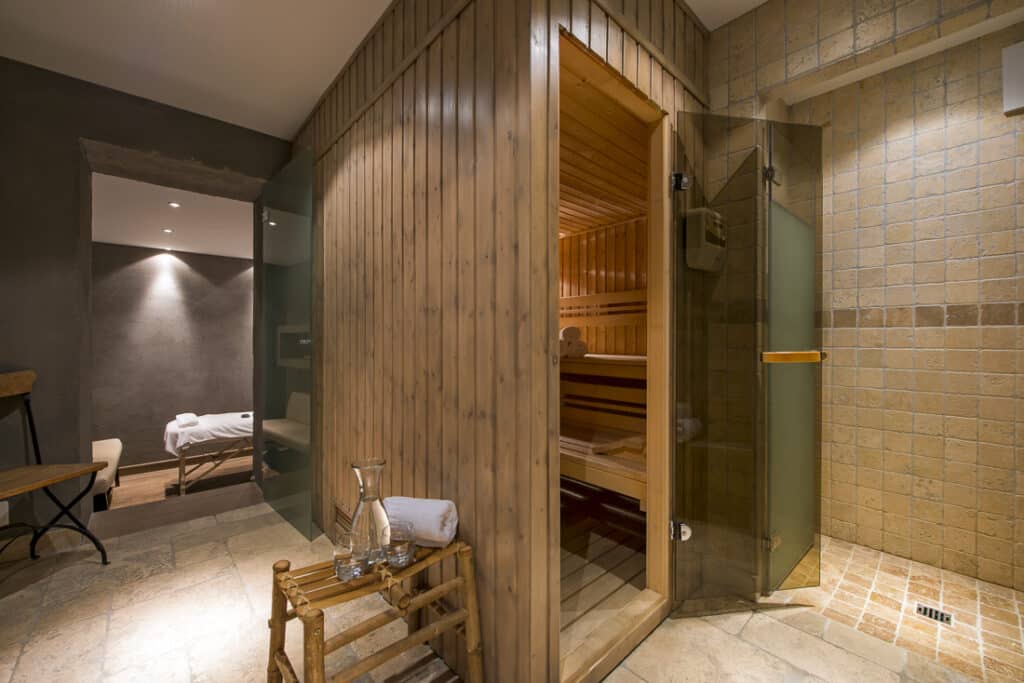 A cozy spa room exudes an air of luxury travel, featuring a wooden sauna with a glass door. Inside, wooden benches invite relaxation. Nearby, a rustic towel rack holds a rolled white towel and a glass jug, while in the dimly lit background rests an inviting massage table. David Pearson Travel