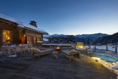At dusk, the alpine cabin terrace offers a luxurious travel experience with its wooden deck, cozy seating, and inviting firepit. Snow blankets the area, while a hot tub with glowing lights adds warmth. The scene is completed by snow-capped mountains and a clear, darkening sky. David Pearson Travel