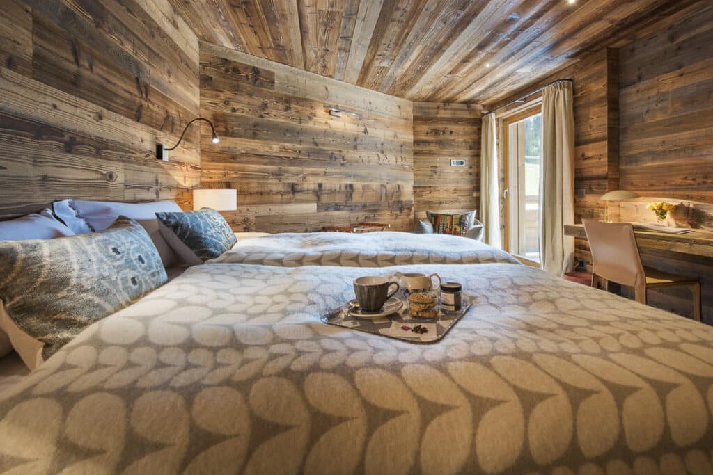 Cozy rustic bedroom with wooden walls and ceiling offers a touch of luxury travel. A plush bed, draped in a patterned blanket, features a breakfast tray with mugs and a jar. Natural light streams through the window, illuminating a desk corner adorned with a lamp and plant. David Pearson Travel