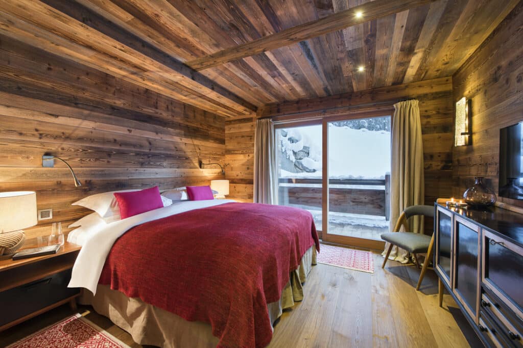 Cozy wooden cabin bedroom, perfect for luxury travel, with a large bed dressed in red blankets and pillows. A window reveals a snowy outdoor scene. Soft lighting, a small chair, and a side table with a lamp complete the warm and inviting atmosphere. David Pearson Travel