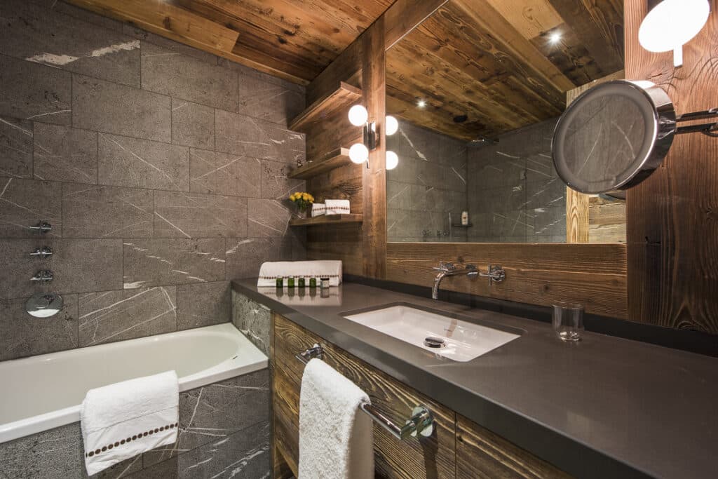 A modern bathroom with a gray stone wall, wooden ceiling and walls exudes luxury travel vibes. It features a bathtub with a towel, a large mirror with lights, a sink with toiletries, and a round magnifying mirror. Shelves adorned with decorative items elevate the ambiance. David Pearson Travel