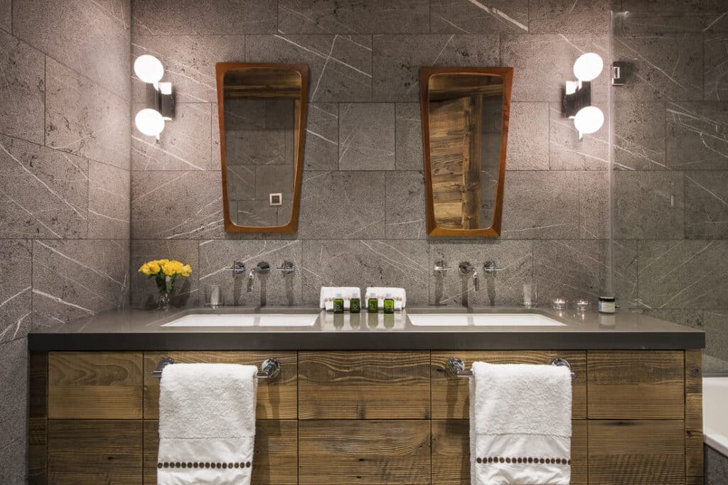 A modern bathroom evokes the elegance of luxury travel with its double sink, wooden framed mirrors, and contemporary light fixtures. The countertop displays toiletries and yellow flowers, while two plush white towels hang from the wooden vanity against a gray textured wall. David Pearson Travel