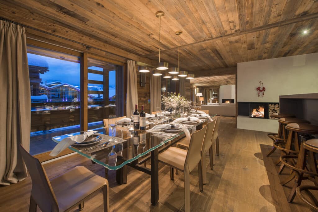 A cozy dining room with a glass table set for a meal, surrounded by wooden chairs, embodies the essence of luxury travel. The room features a wooden ceiling and walls, a modern fireplace with a mantle, and large windows revealing a scenic evening view. Warm lighting creates an inviting atmosphere. David Pearson Travel
