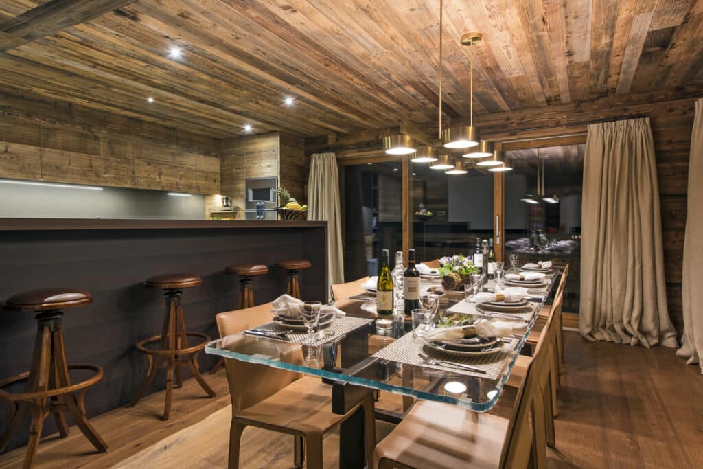 A cozy dining area with a wooden ceiling and walls exudes the charm of luxury travel. A glass table set with white plates, glasses, and napkins is surrounded by six tan chairs. In the background, a kitchen counter with stools awaits, while warm pendant lights cast an inviting glow. David Pearson Travel