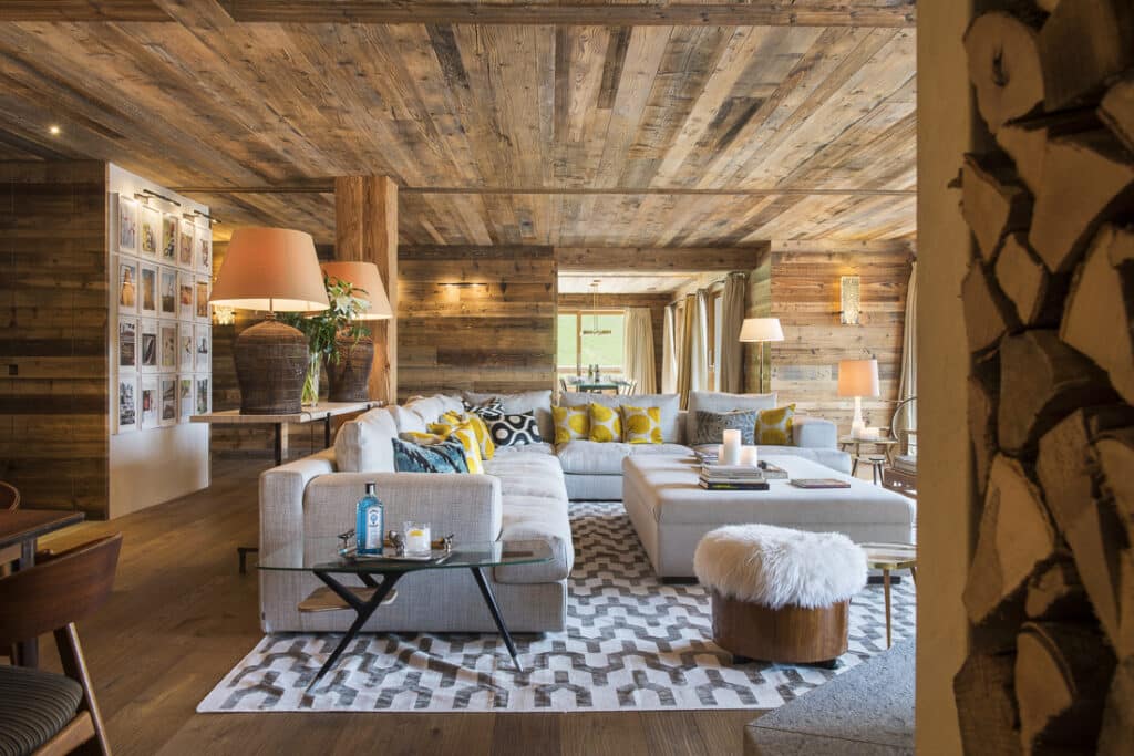 A cozy living room exuding the charm of luxury travel, with a rustic wooden ceiling and walls. It features a large beige sectional sofa adorned with yellow and patterned cushions, a chic rug, a small glass coffee table, and various lamps. A stack of firewood adds to the sophisticated ambiance. David Pearson Travel