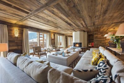 Spacious living room with wooden walls and ceiling, featuring a luxury travel-inspired large sectional sofa with colorful cushions, a central coffee table, and a fireplace. Large windows offer a view of mountains, and natural light fills the room. David Pearson Travel