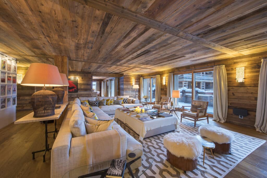 The spacious living room exudes luxury travel vibes with its wooden walls and ceiling, featuring a large beige sofa, patterned rug, coffee table with books, two fluffy stools, and expansive windows showcasing a snowy exterior. Warm lighting and modern decor create a cozy atmosphere. David Pearson Travel