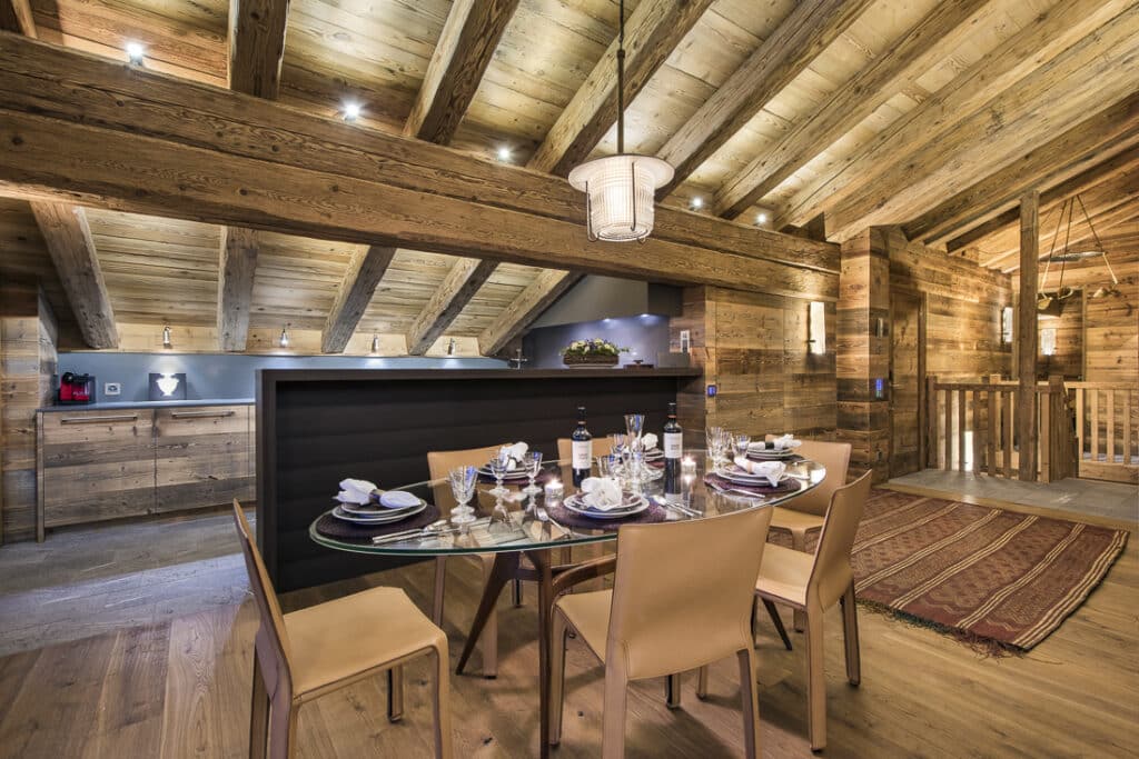 A cozy dining area with a glass table set for six, tan chairs, and rustic wood-paneled walls and ceiling. Warm lighting highlights the wooden beams and an open kitchen in the background. David Pearson Travel