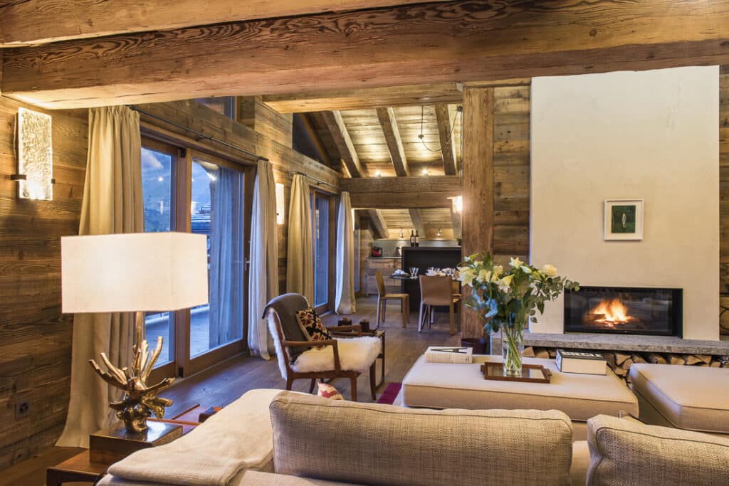 Cozy living room with rustic wooden beams, beige sofas, a lit modern fireplace, and a vase of flowers on a coffee table. Large windows with curtains reveal an outdoor view reminiscent of luxury travel destinations. Dining area visible in the background under warm lighting. David Pearson Travel