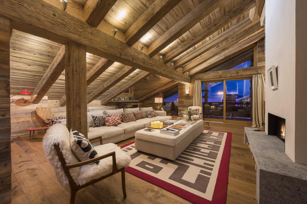 Cozy living room with exposed wooden beams and a sloped ceiling, offering a luxury travel vibe. Features a large sectional sofa with patterned pillows, a coffee table on a geometric rug, and a modern fireplace. Sliding glass doors provide an exquisite view of the evening sky. David Pearson Travel