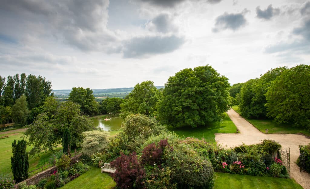 A vast, lush garden with a gravel path leading through it provides an exquisite escape reminiscent of luxury travel. Trees and bushes surround the area, with a pond visible in the distance. The sky is cloudy, adding a dramatic feel to the landscape. David Pearson Travel