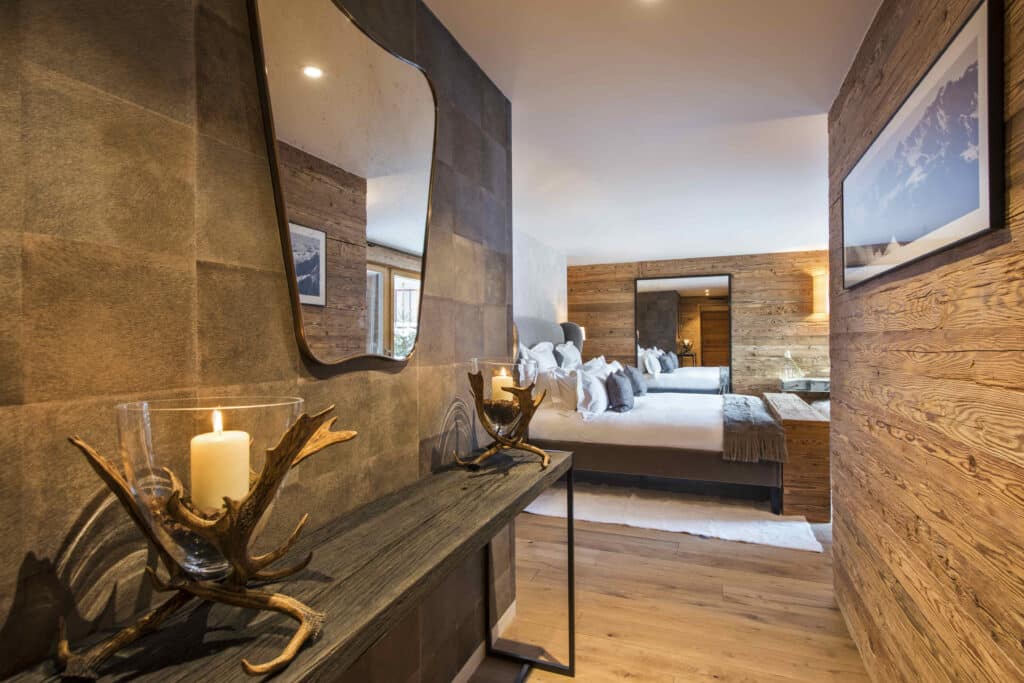 A cozy, modern bedroom perfect for luxury travel enthusiasts, featuring a warm, rustic design. A wooden wall holds a framed picture, while a console with antler candle holders stands in the foreground. The large bed, adorned with plush pillows and cushions, invites relaxation and indulgence. David Pearson Travel