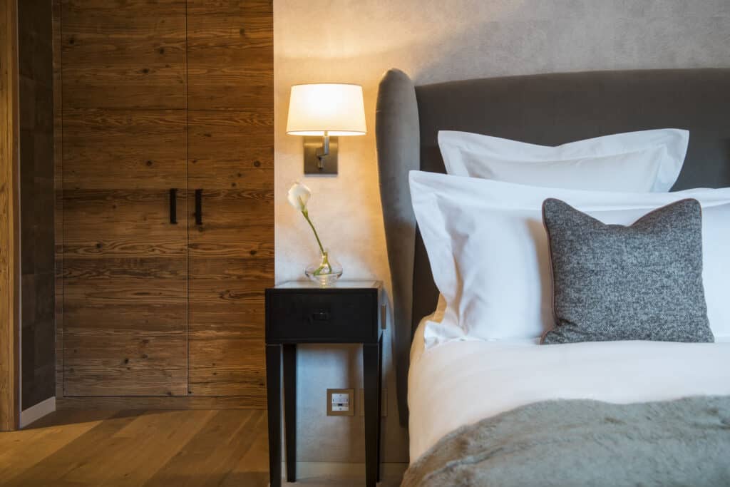 A cozy bedroom exudes luxury travel vibes, featuring a large bed with white linens and a gray pillow. A small black nightstand beside the bed holds a single flower in a vase. A warm wall lamp is lit, and a wooden closet with double doors stands elegantly in the background. David Pearson Travel