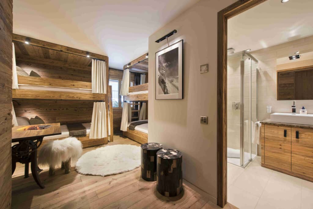 Cozy room with wooden bunk beds and a fluffy white rug on wooden floors, offering a hint of luxury travel. A small table with a fur-covered stool sits by the entrance. Adjacent is a bathroom featuring a glass-enclosed shower and sleek wooden cabinetry. A framed picture adorns the wall. David Pearson Travel