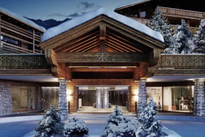 A luxurious mountain hotel entrance, epitomizing luxury travel, features a wooden and stone facade adorned with snow. The building glows warmly beneath a snowy blanket, encircled by snow-laden pine trees, crafting a cozy winter retreat. David Pearson Travel