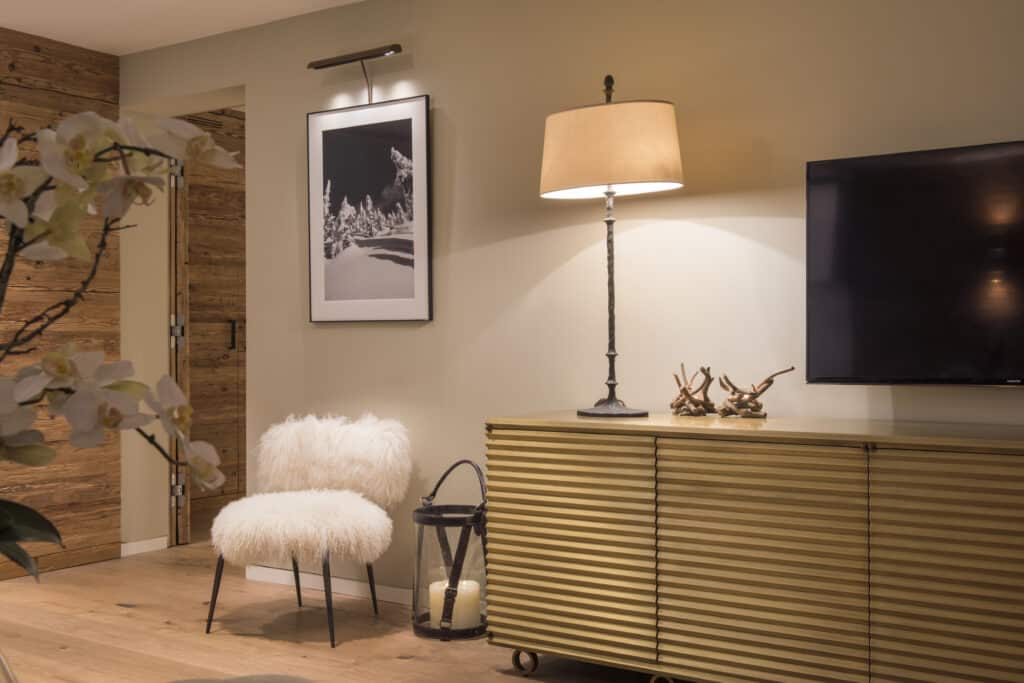 Stylish living room with a cozy sheepskin chair, a modern lamp on a corrugated sideboard, and a large black-framed photo that evokes luxury travel. A lantern with candles sits on the floor, casting an inviting glow reminiscent of an exotic getaway. A TV is mounted on the right wall. David Pearson Travel