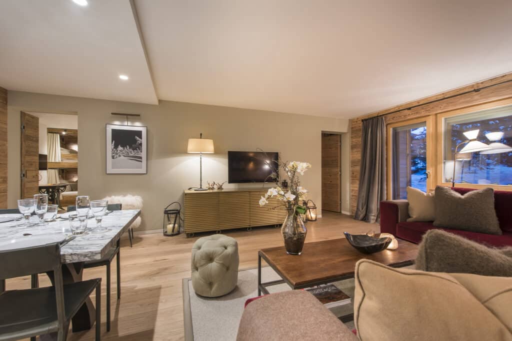 Cozy living room with a modern design reminiscent of luxury travel, featuring a dining area with elegant table settings, a plush maroon couch adorned with pillows, a wooden TV cabinet, and decorative plants. Large windows invite natural light while neutral tones create a warm atmosphere. David Pearson Travel