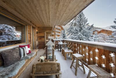 A cozy wooden balcony with rustic seating offers a luxury travel experience, overlooking a snowy landscape with evergreen trees and wooden chalets. The railing, adorned with star cut-outs, complements the fluffy cushions and blankets arranged on the bench. David Pearson Travel
