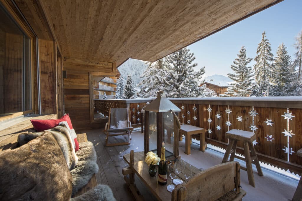 A luxury travel escape: a wooden balcony overlooking a snowy landscape, adorned with star ornaments. Cozy seating with fur cushions beside a small table featuring champagne, glasses, and flowers. Pine trees and snow-covered mountains complete this idyllic scene. David Pearson Travel