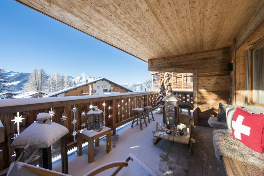 Cozy wooden chalet balcony overlooking snow-covered mountains and trees. This luxury travel retreat features cushioned seating, lanterns, and a small table adorned with candles and snow, crafting a warm and inviting winter scene. David Pearson Travel