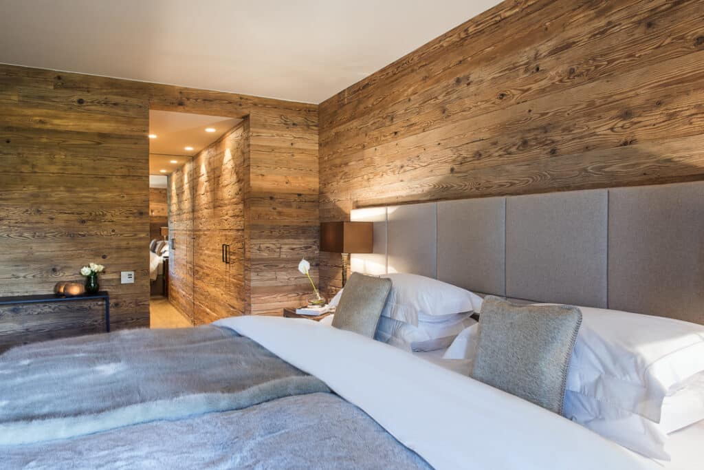 A cozy bedroom exuding luxury travel vibes features a large bed with gray and white linens, wooden walls, and soft lighting. A pathway leads to a doorway. A small lamp and decorative items grace the side table, enhancing the warm and inviting atmosphere. David Pearson Travel