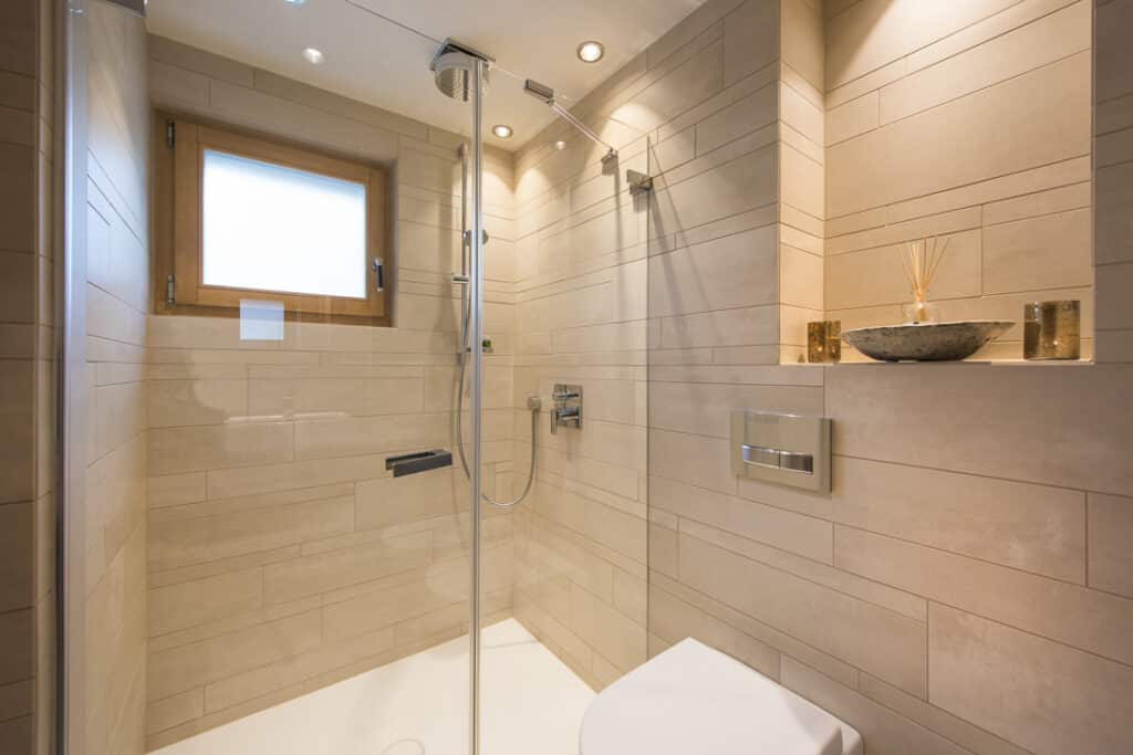 Experience a touch of luxury travel in a modern bathroom featuring beige tiled walls and a glass-enclosed shower with an overhead rain showerhead. Natural light filters through a small square window, while elegant touches include a decorative bowl with reeds on the shelf. David Pearson Travel