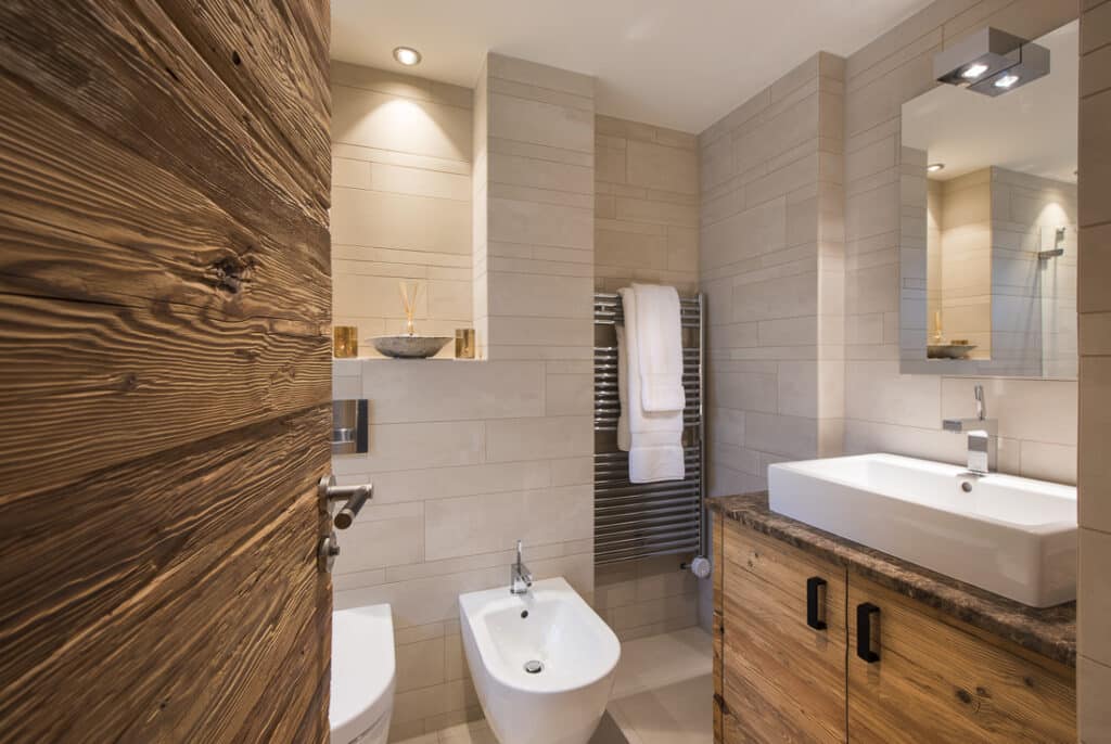 A modern bathroom with a wooden door, white toilet, bidet, and wooden vanity evokes the essence of luxury travel. The vanity features a white sink, large mirror, and overhead light. Beige tiles cover the walls while a towel hangs on a metal rack. Warm lighting creates a cozy atmosphere. David Pearson Travel