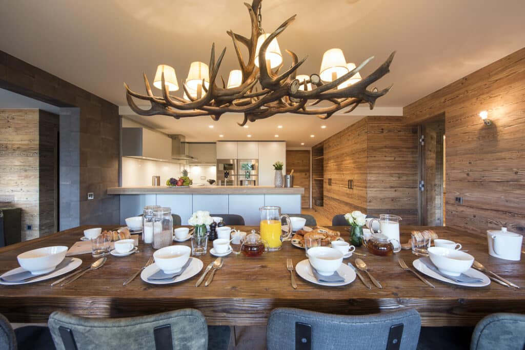 A rustic dining room with a large wooden table set for breakfast evokes the charm of luxury travel. Plates, cups, and glasses are arranged neatly with orange juice and pastries. A unique antler chandelier hangs overhead, while wooden walls frame a modern kitchen in the background. David Pearson Travel