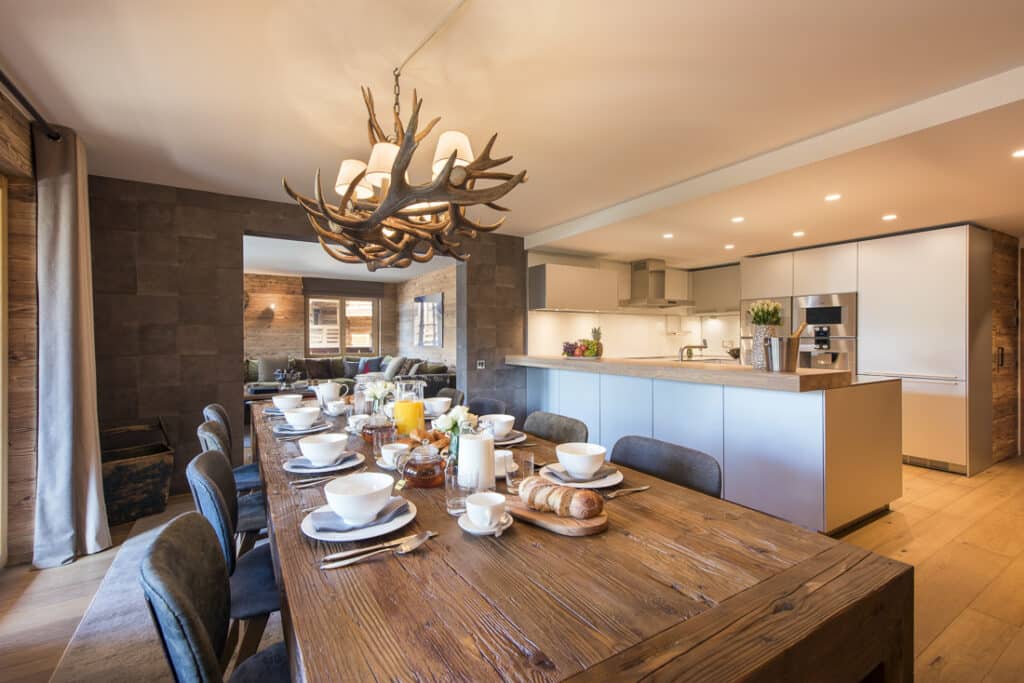 The rustic dining room, perfect for a luxury travel retreat, features a long wooden table set for breakfast. Tableware includes plates, cups, and fresh bread beneath an antler chandelier. The open kitchen in the background is modern with white cabinets and stainless steel appliances. David Pearson Travel