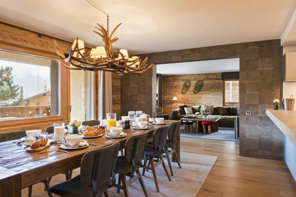 A dining room with a large wooden table set for breakfast, featuring pastries and white dishes, exudes luxury travel vibes. An antler chandelier hangs above. The room opens into a cozy living area with a sofa and rustic decor. Large windows offer natural light and a view outside. David Pearson Travel