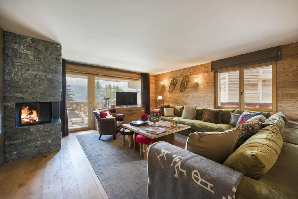 A cozy living room reminiscent of luxury travel features a stone fireplace, wooden walls, and large windows with scenic views of trees and mountains. The space boasts a large green sectional sofa with cushions, a wooden coffee table, and a TV on a stand for ultimate relaxation. David Pearson Travel