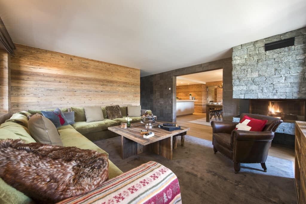 A cozy living room reminiscent of luxury travel, featuring a large green sectional sofa with colorful cushions, a wooden coffee table, and an armchair adorned with a red pillow. A stone fireplace graces the right side, while an elegant dining area is visible through an open doorway. David Pearson Travel
