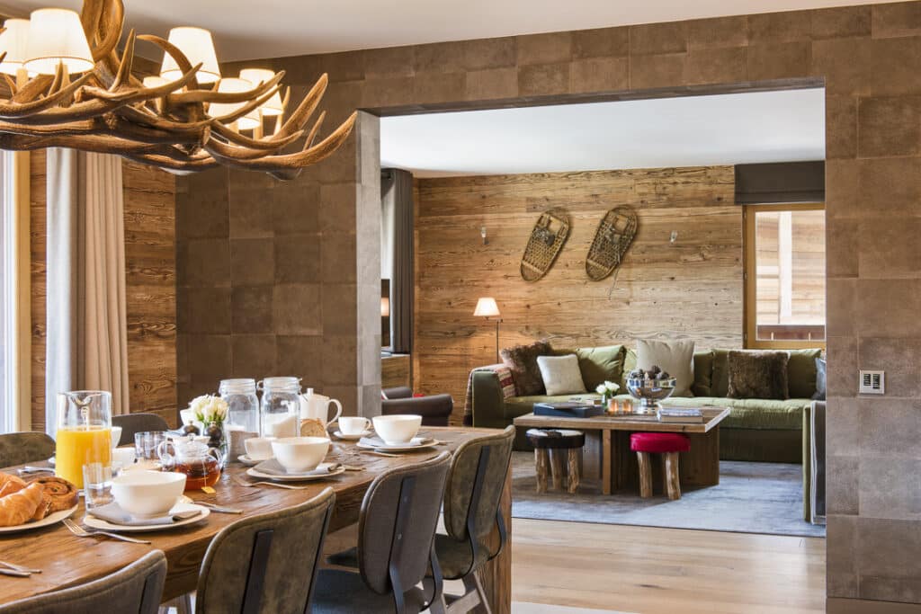 A cozy dining area with a wooden table set for a luxurious breakfast, featuring chairs and a rustic chandelier. Beyond is a living room with a green sofa, wooden walls, and snowshoes as decor. Natural light streams through the window, creating an inviting luxury travel ambiance. David Pearson Travel