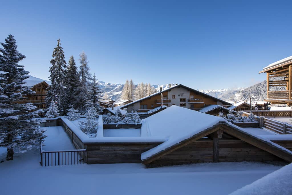 Snow-covered wooden cabins and rooftops in a mountain village offer a glimpse of luxury travel amidst nature. Surrounding trees and distant mountains enhance the serene winter scene under a clear blue sky. David Pearson Travel